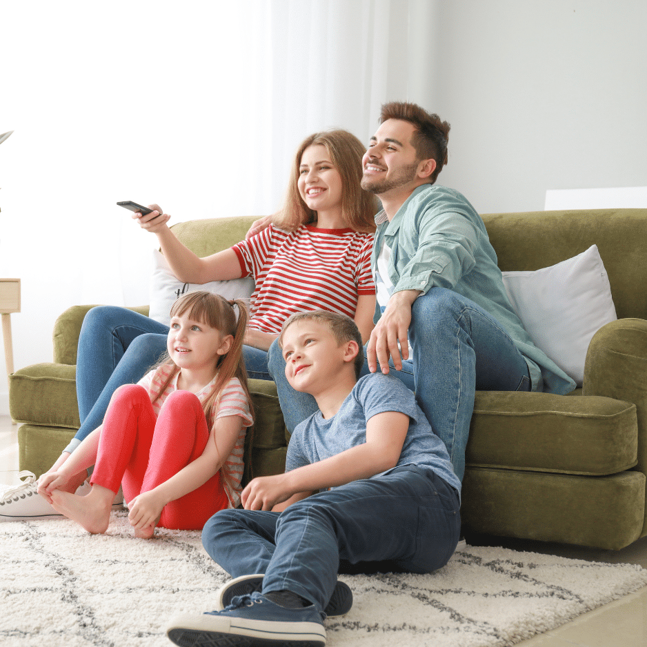 A family of four sits together in a cozy Sunshine Coast living room. The parents are on a green couch; the mother holds a remote control, while the father sits to her right. Two children, a girl and a boy, sit on the floor in front of them, all smiling and looking at a TV.