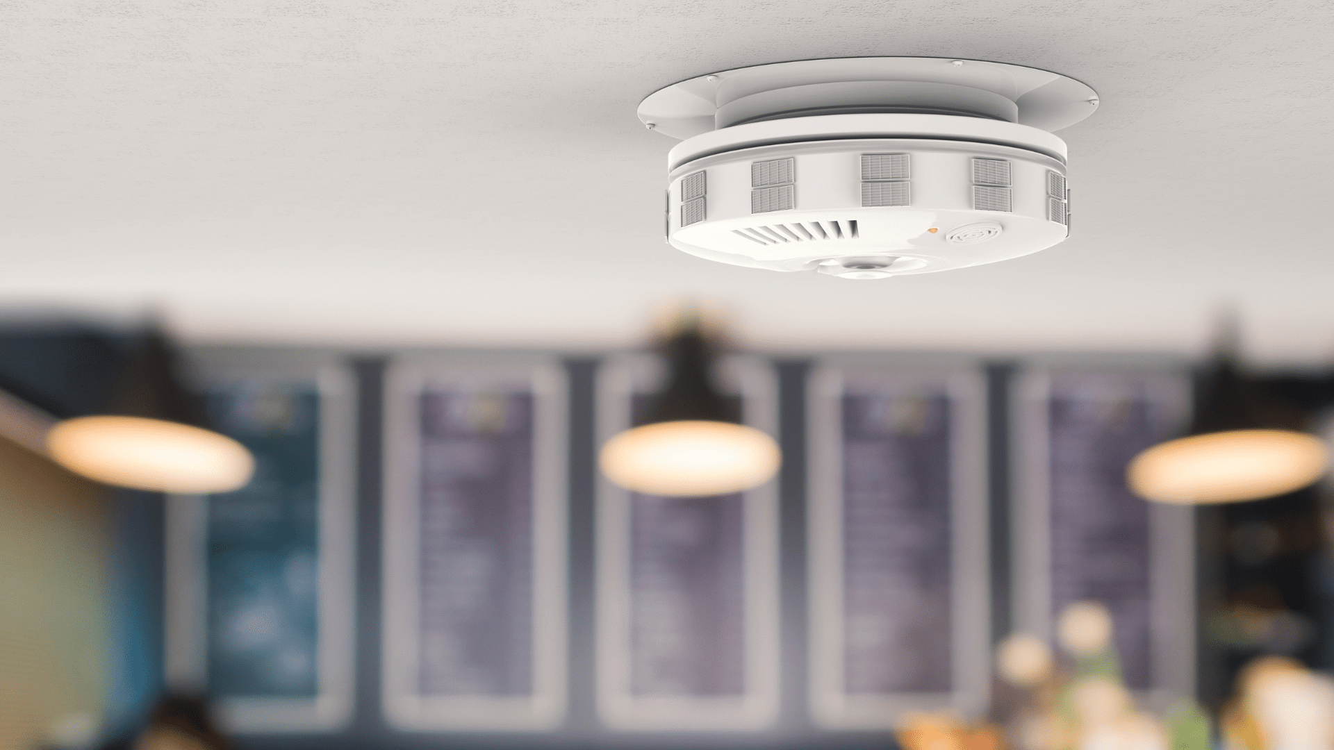 A smoke detector is mounted on the ceiling of a room, possibly somewhere like a quaint café on the Sunshine Coast. The background is blurred, with hanging lights and what appear to be large menu boards on the wall, adding to the warm and inviting atmosphere.