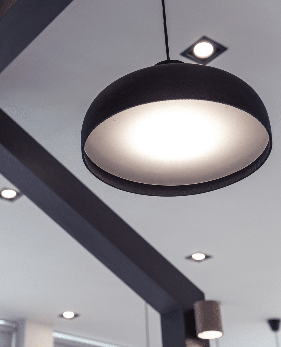 A modern hanging pendant light fixture is illuminated, casting a soft glow reminiscent of the Sunshine Coast. The light has a black exterior and a white interior, suspended from the ceiling by a cord. Several recessed ceiling lights are also visible in the background.
