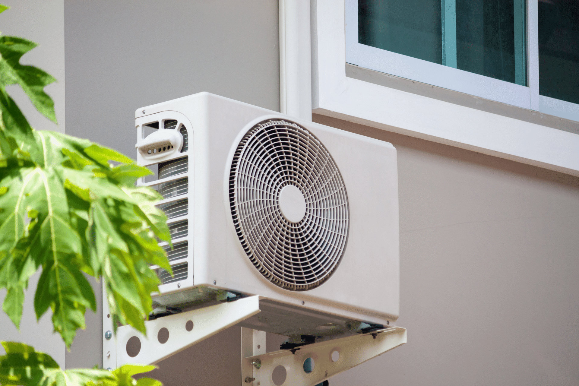 A white outdoor air conditioning unit is mounted on the exterior wall of a building near a window, with green leaves partially obscuring the view. Situated on the Sunshine Coast, this serene setup blends modern convenience with natural beauty.