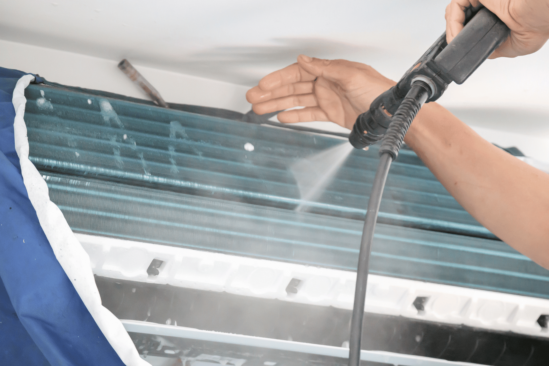 A person is cleaning an air conditioning unit using a high-pressure water spray on the Sunshine Coast. The individual is holding the spray nozzle with one hand and meticulously cleaning the metal fins and coils of the unit. Part of a blue protective cover and a white wall are visible in the background.