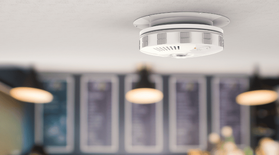 A smoke detector is mounted on a ceiling in an indoor space. There are blurred lights and posters in the background, hinting at the vibrant life of the Sunshine Coast. The smoke detector is round and white with small vents and a central sensor, suggesting this could be a public or commercial area.