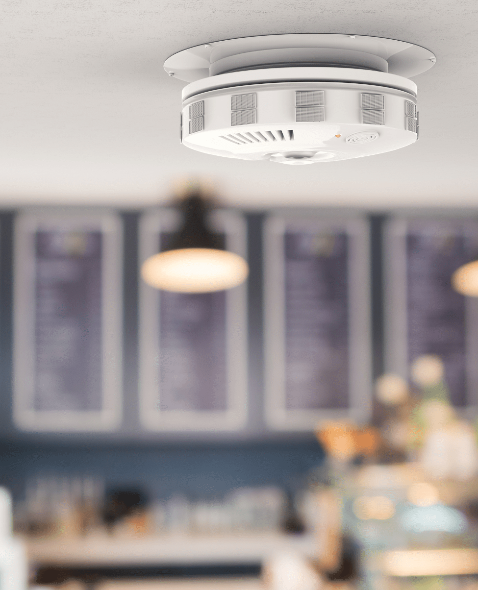 A smoke detector is mounted on the ceiling of a well-lit room, with blurred menus and hanging light fixtures visible in the background. The room exudes a modern and inviting ambiance, reminiscent of the relaxed atmosphere found on the Sunshine Coast.