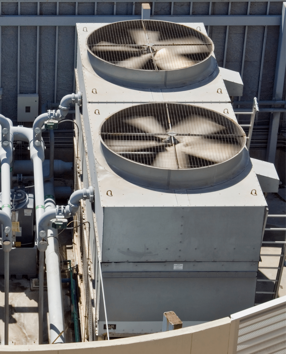 A top view of two large industrial fans housed in metal casings on a building's rooftop along the Sunshine Coast. Several pipes and valves are visible around the fans, indicating a complex mechanical setup, likely part of an HVAC system.