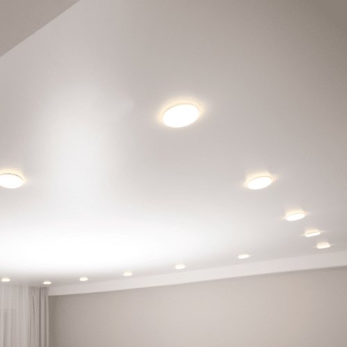 A modern ceiling with multiple small, round recessed lights. The lights are uniformly distributed, providing a soft, even illumination reminiscent of the Sunshine Coast. White walls complete the minimalist design, and sheer curtains are visible in the corner.