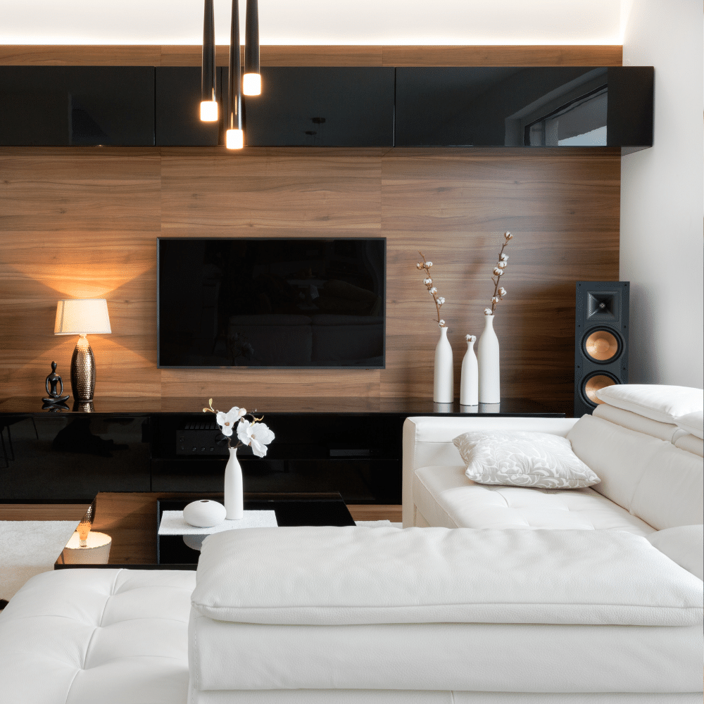 A modern living room with wood-paneled walls, a black flat-screen TV, and black and wooden storage units evokes the laid-back elegance of the Sunshine Coast. White sofas, cushions, a small side lamp, and decorative vases adorn the space. Multiple hanging lights illuminate the room.