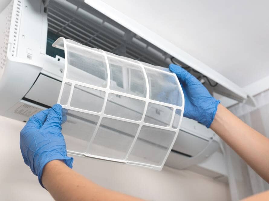 A person wearing blue gloves holds up a clean air filter, about to insert it into an open air conditioning unit mounted on a wall. The person's hands and forearms are visible, while the internal components of the air conditioner are exposed. It's a typical maintenance task on the Sunshine Coast.