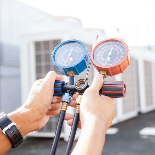 Person's hands holding a manifold gauge set with blue and red dials, used for HVAC systems, under the bright skies of the Sunshine Coast. Industrial air conditioning units loom in the background as the gauges check pressures, indicating maintenance or repair work.