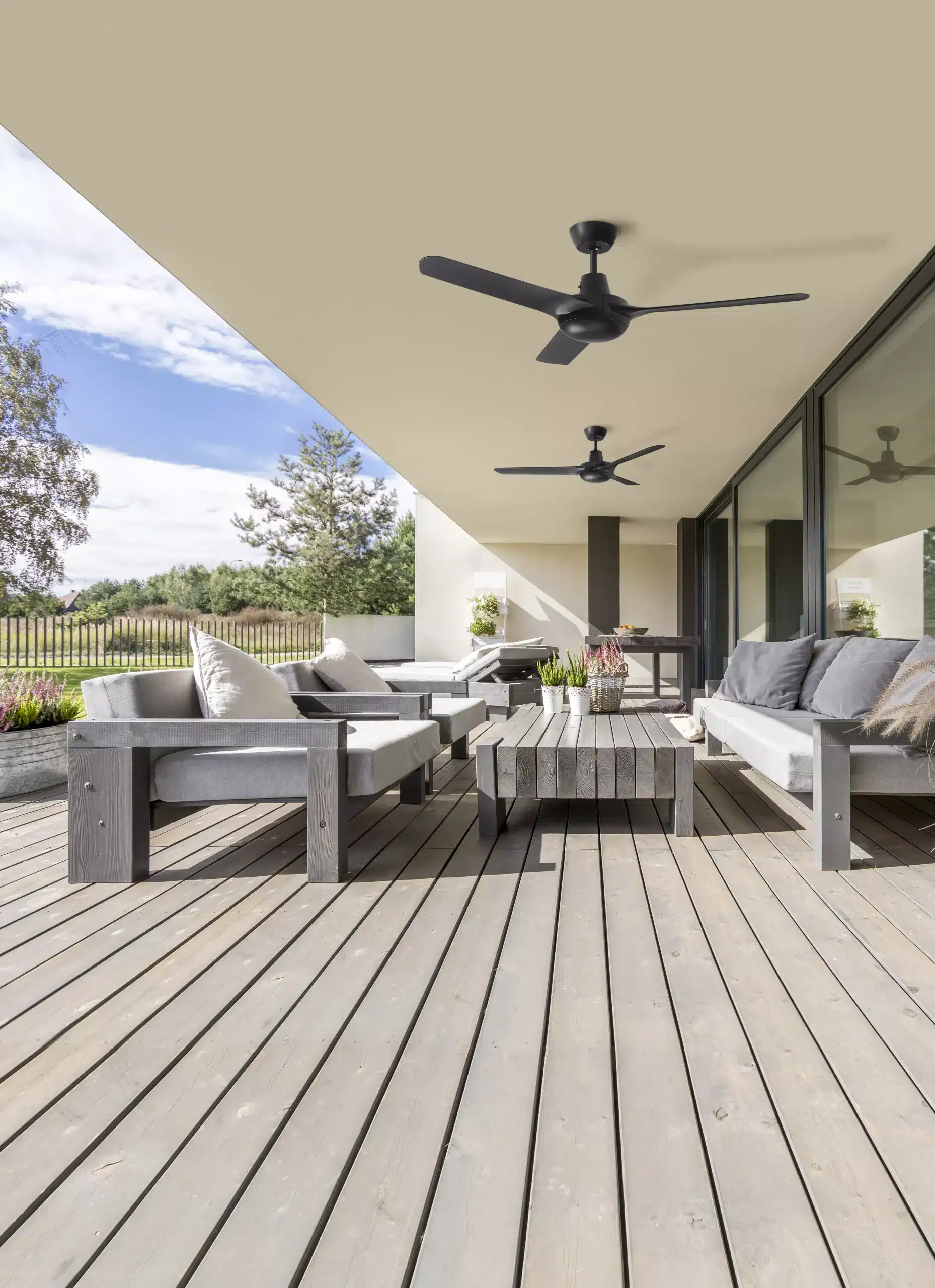 A modern outdoor patio with light wood decking, perfect for enjoying the Sunshine Coast. It features two ceiling fans, grey cushioned sofas, a wooden coffee table, and potted plants. Large glass doors lead inside, and a fence and trees are visible in the background under a blue sky.