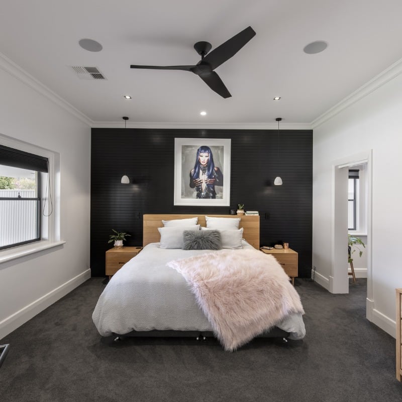 A modern bedroom reminiscent of sunshine coast vibes features a central bed with white linens and a pink furry throw, wooden nightstands, and wall-mounted lamps against a dark accent wall. A ceiling fan is above the bed, while large windows and doorways invite ample natural light.