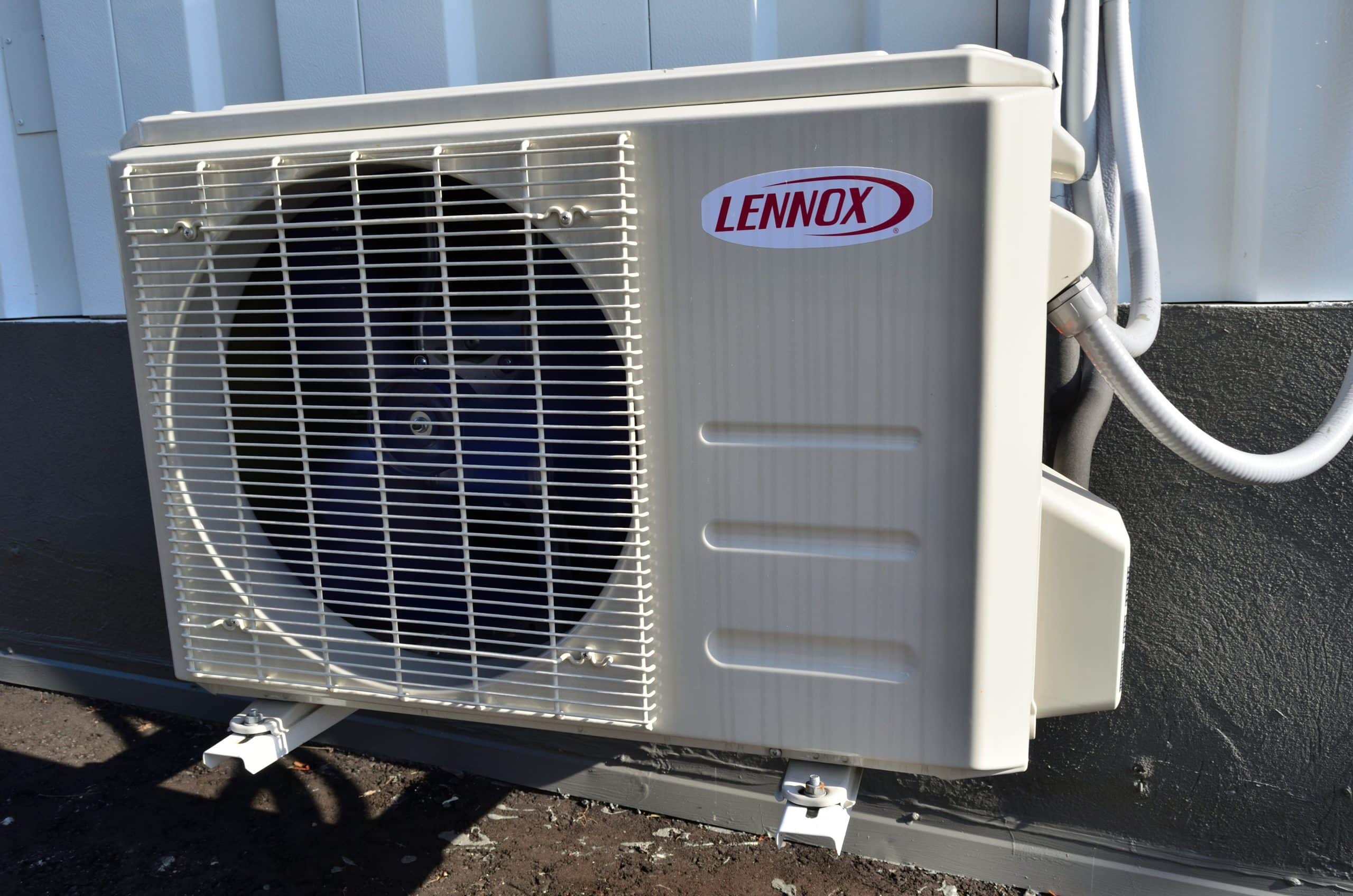 A Lennox outdoor air conditioning unit is mounted against a building’s exterior wall, with visible vents, pipes, and wiring connected to it.