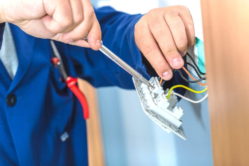A person wearing a blue jacket uses a screwdriver to install or repair electrical wiring in a wall outlet. Several colored wires are visible, and pliers hang from their pocket.