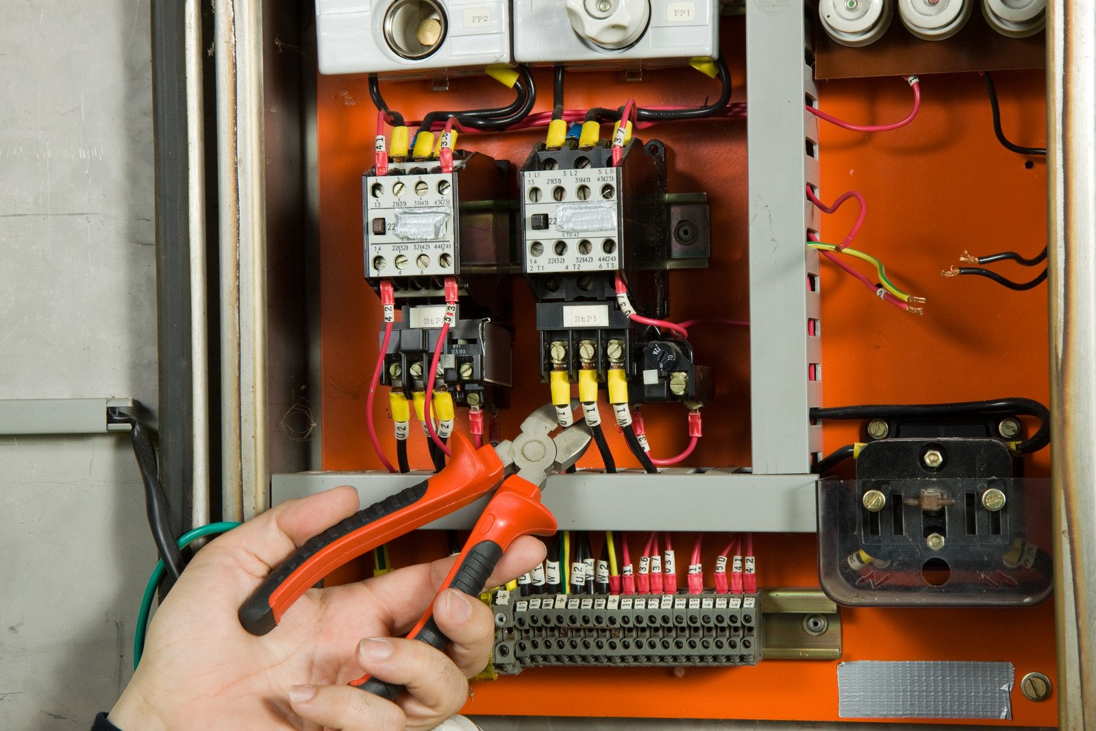 A hand holding red-handled wire cutters is positioned in front of an open electrical control panel with exposed wires, switches, and circuit components.