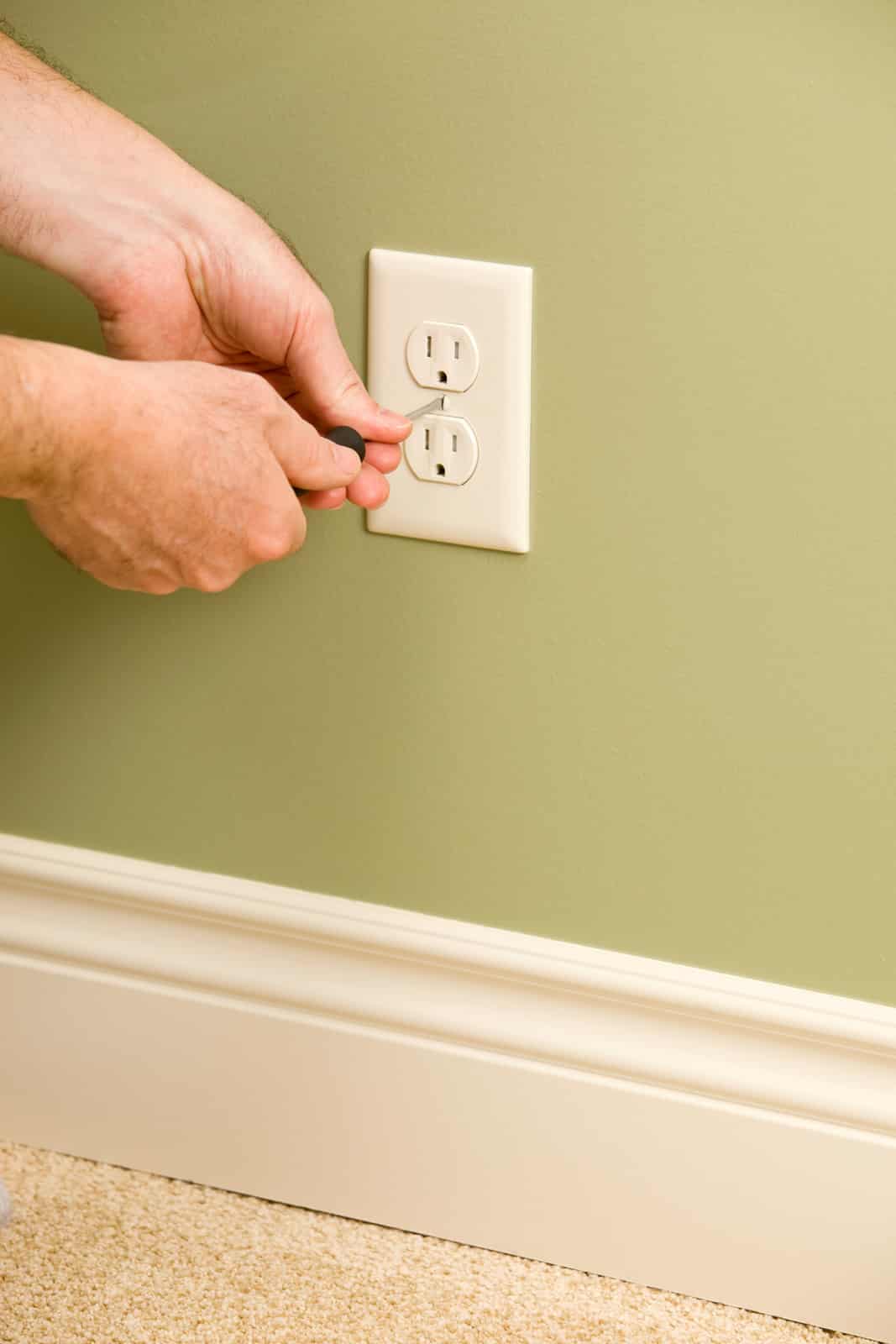 A person uses a screwdriver to remove the cover plate from a white electrical outlet on a green wall above a beige carpeted floor with white trim.